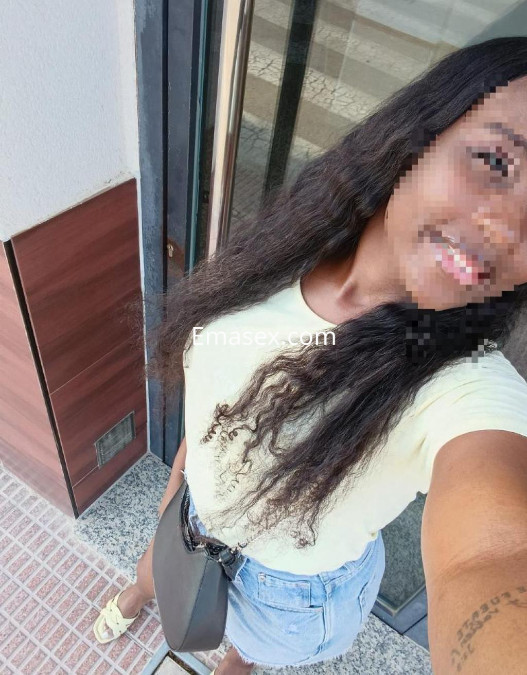 Escort en Murcia con cabello largo y rizado, vistiendo una camiseta amarilla y shorts de mezclilla, sonriente en la calle.