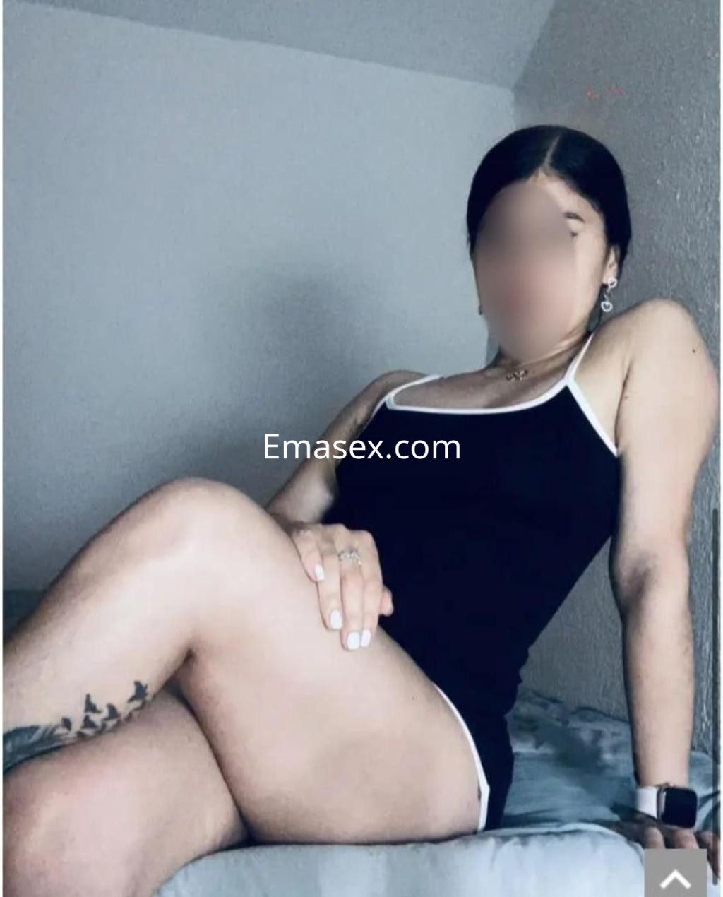 Escort en Huelva sentada sobre una cama, vistiendo un top negro sin mangas y con una pierna cruzada, mostrando un tatuaje.