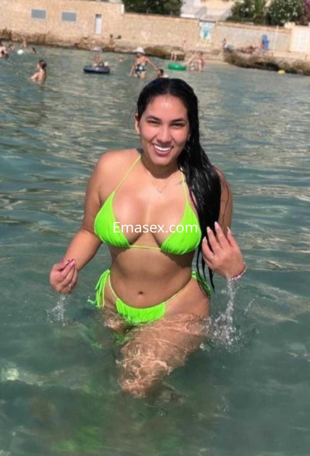 Puta en Huelva luciendo un bikini verde neón mientras se refresca en aguas claras, sonriendo felizmente.