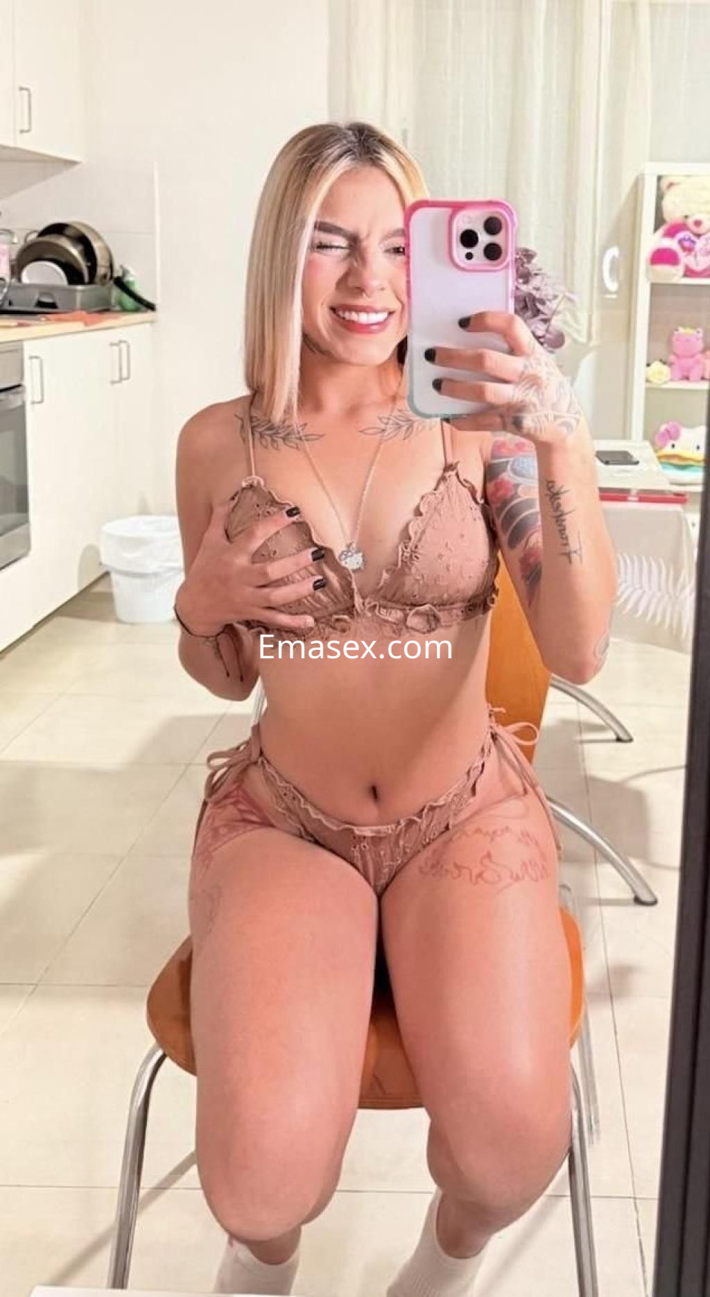 Escort en Murcia sonriente, posando en lencería color nude, sentada en una silla con un ambiente hogareño.