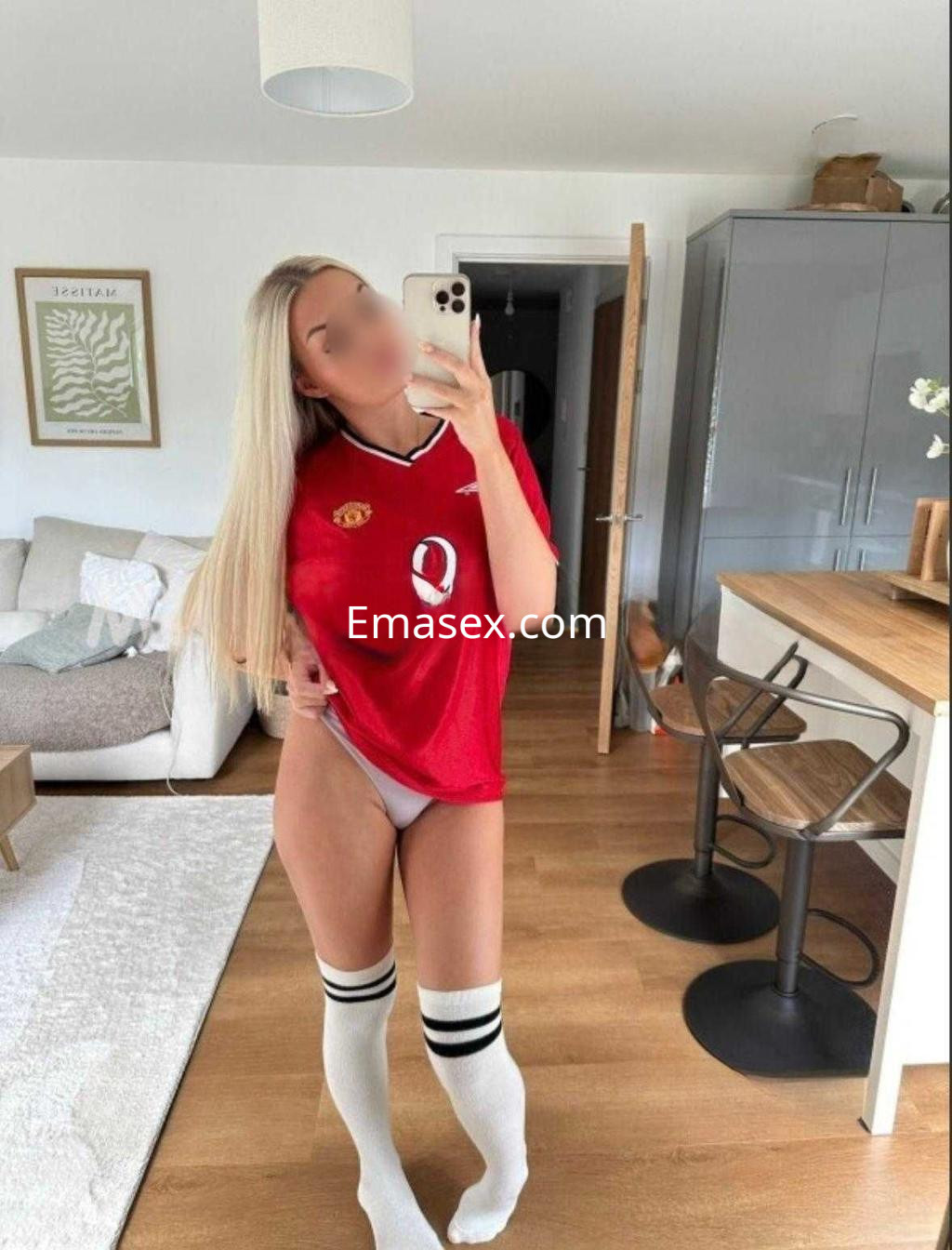 Escort en Barcelona, luciendo una camiseta roja de fútbol y medias largas blancas con rayas en un ambiente moderno.