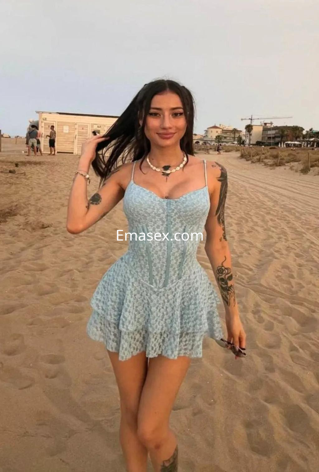 Escort en Palma de Mallorca luciendo un vestido azul claro y mostrando tatuajes, posando en la playa.