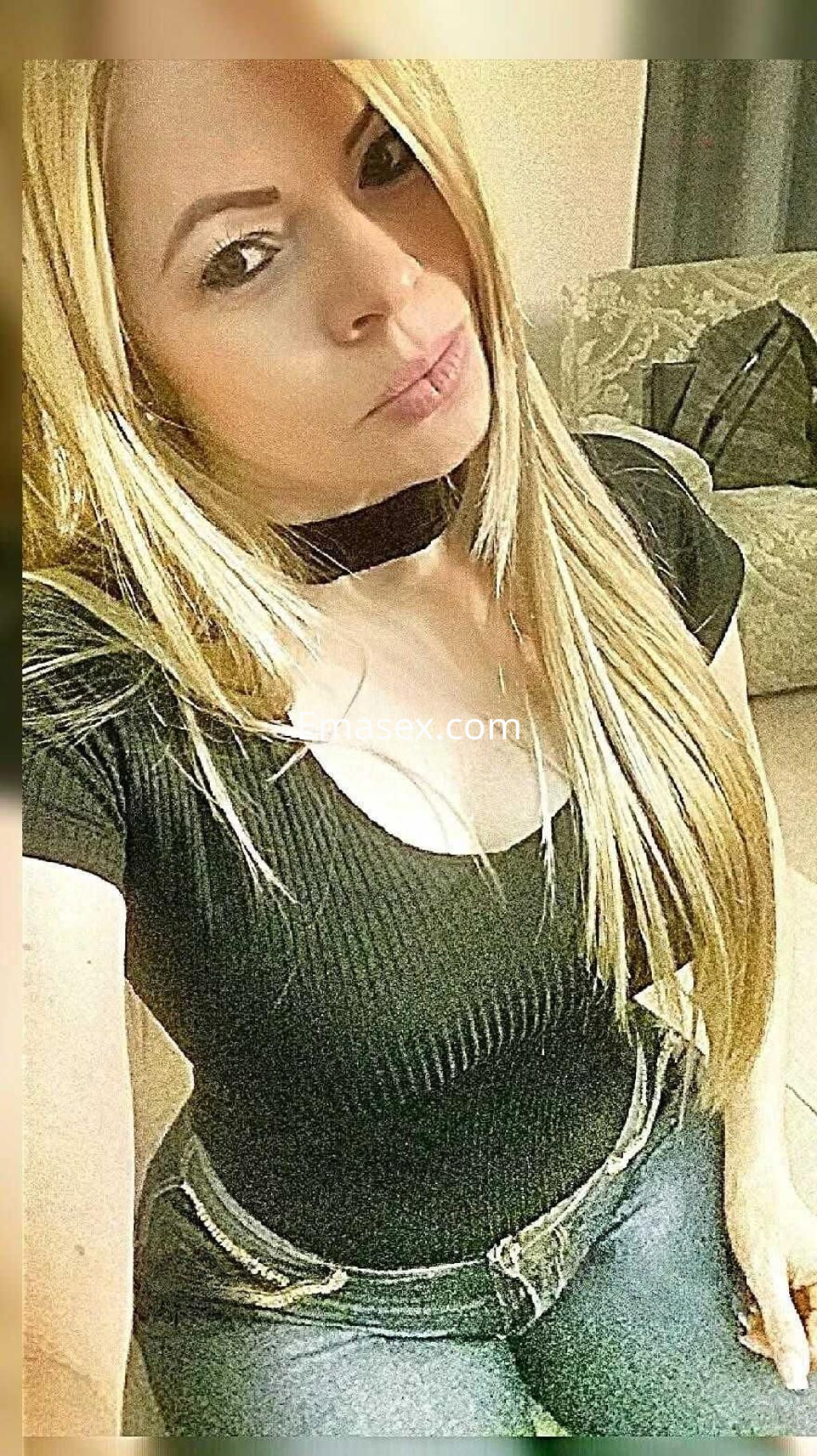 Escort en Madrid, con cabello rubio largo y lacio, viste una blusa negra y jeans ajustados, posando con un leve gesto.