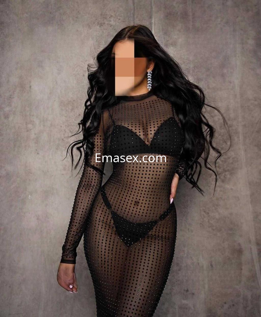 Escort en Barcelona, con cabello largo y rizado, viste un vestido negro transparente con detalles brillantes sobre un fondo neutro.