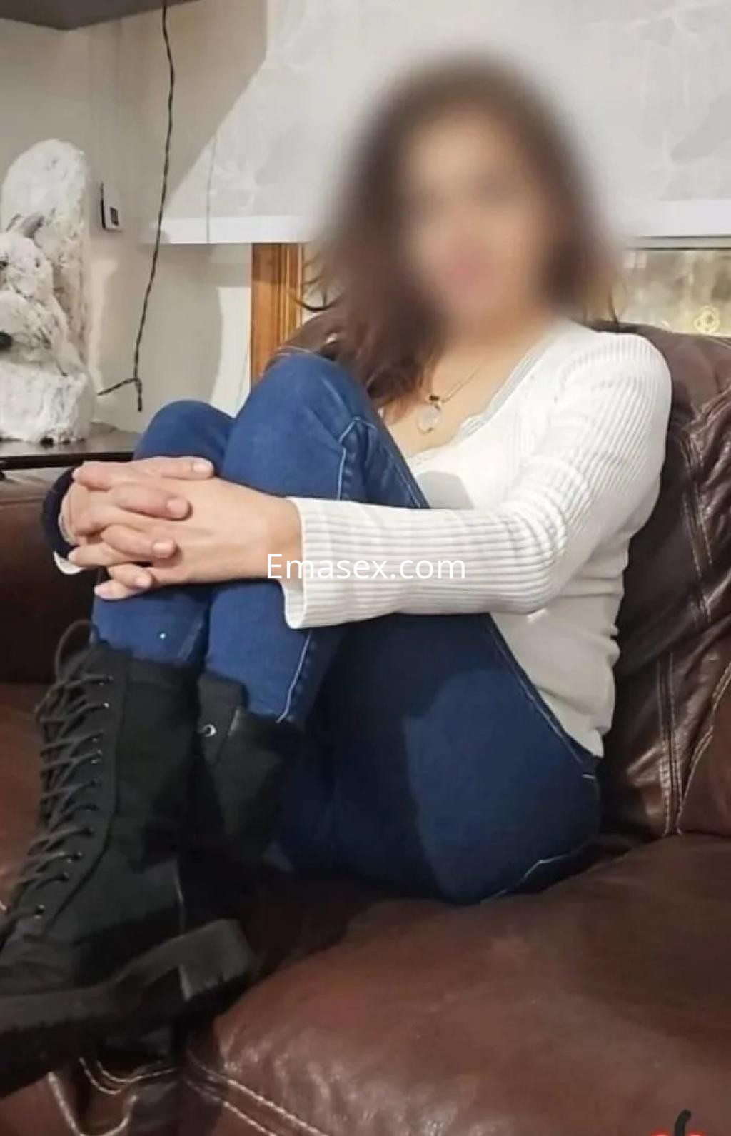 Puta sentada en un sofá, con jersey de manga larga blanco, jeans ajustados y botas negras, en Barcelona.