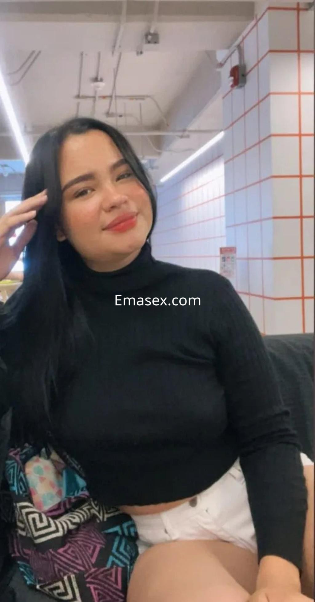 Escort en Zaragoza con blusa negra ajustada y shorts blancos, sonriendo en un ambiente moderno.