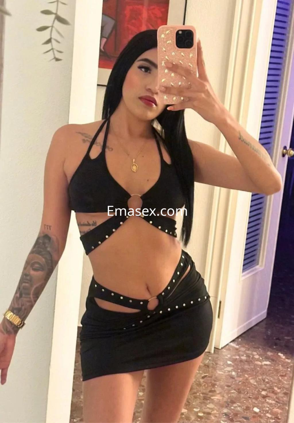 Escort en Palma de Mallorca con cabello largo y lacio, viste un conjunto negro con detalles metálicos y muestra un tatuaje en el brazo.