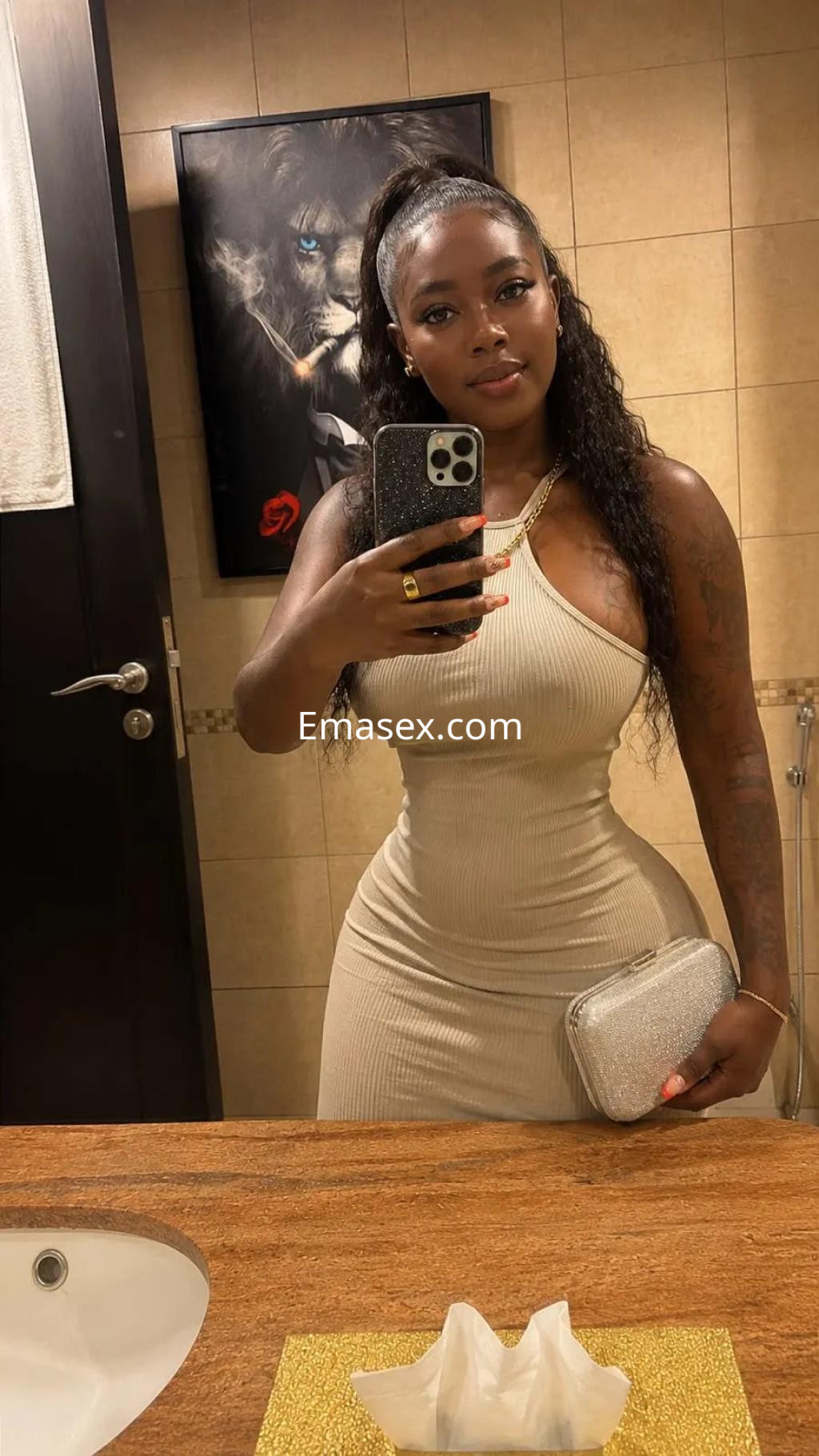 Escort en Zaragoza posando en un baño con un vestido ajustado color beige y bolso plateado, sonriendo a la cámara.