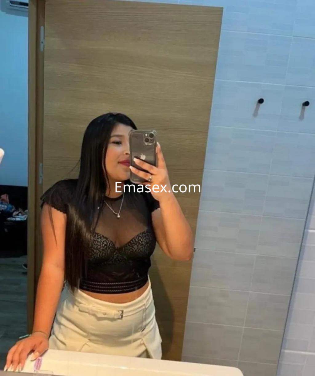 La puta posa frente al espejo en un baño, vistiendo un top negro de encaje y una falda clara, con cabello largo.