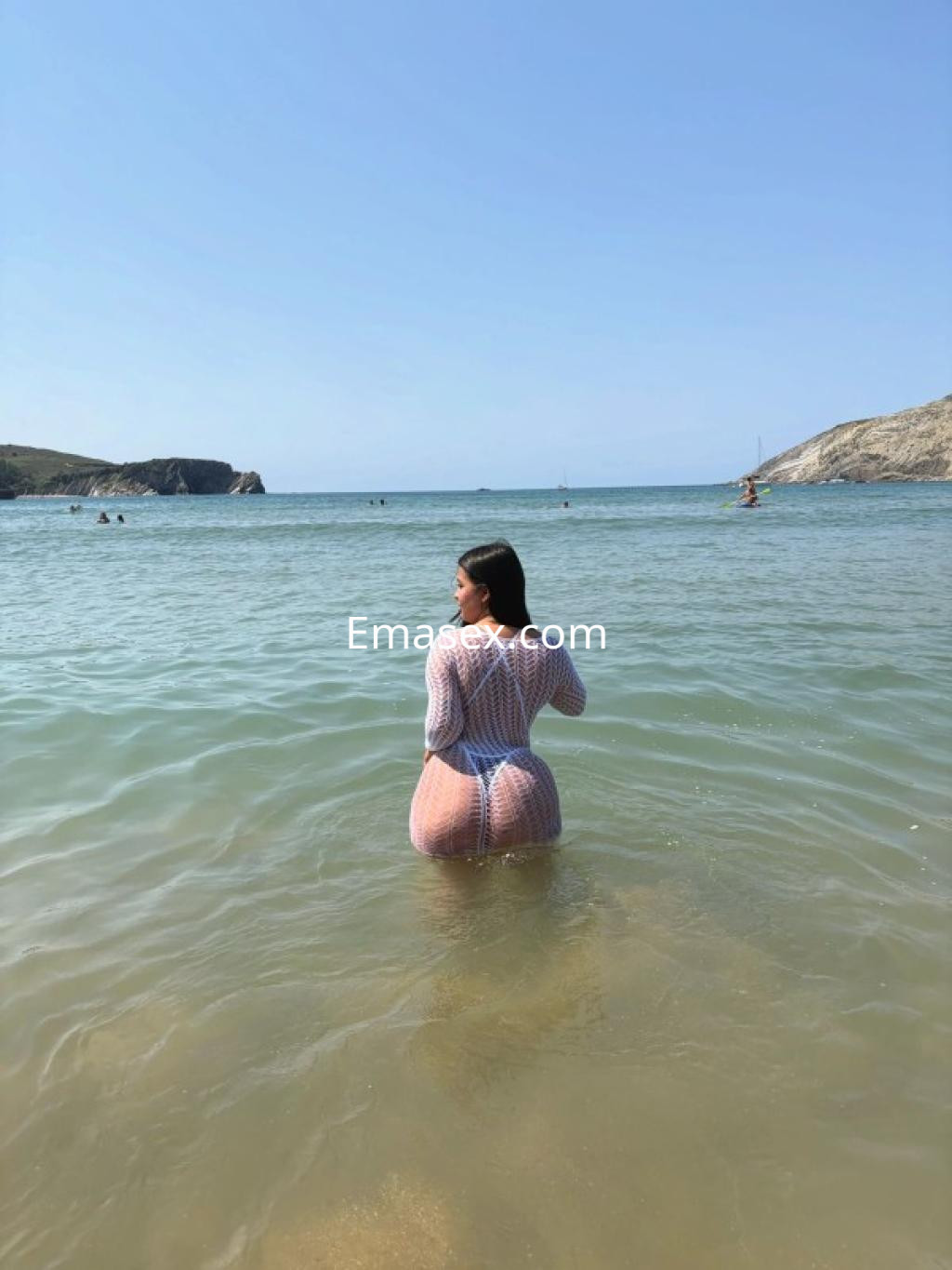 Escort en Ourense con un vestido de malla transparente, posando en el agua de la playa bajo un cielo despejado.