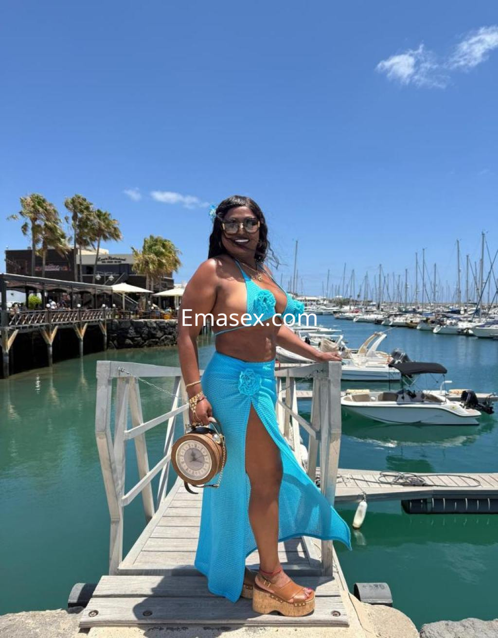 Escort en Las Palmas luciendo un bikini azul y una falda transparente, posando junto al muelle y los barcos.