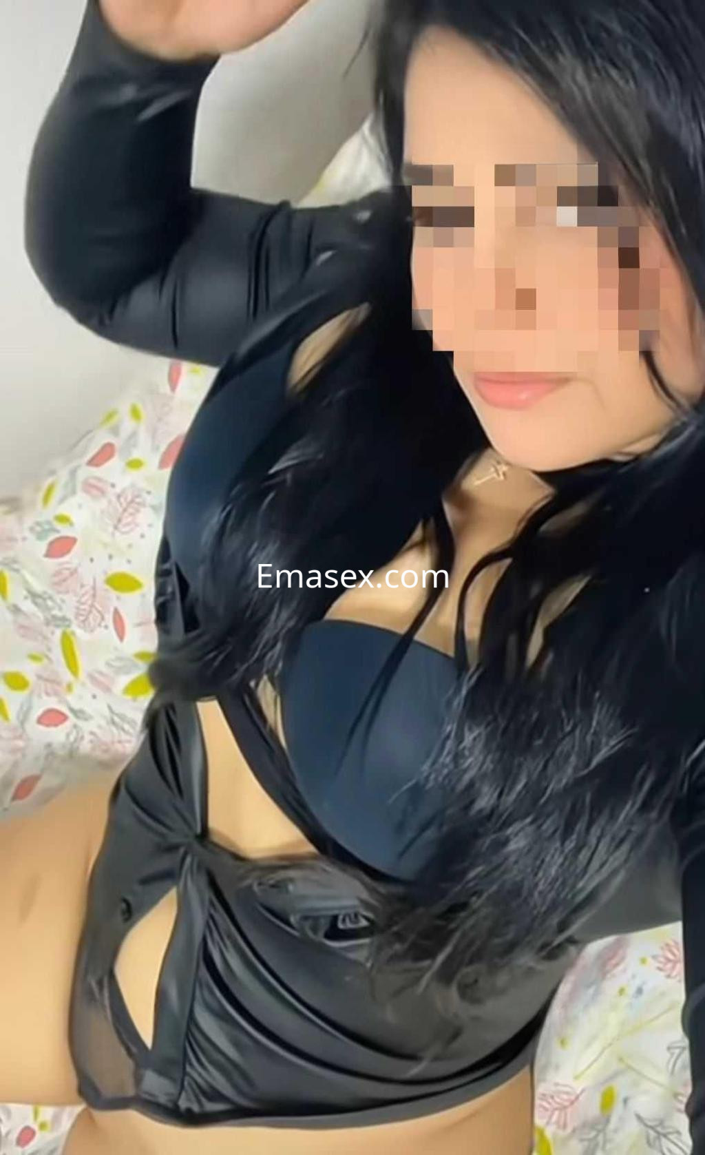 Puta en Huelva luciendo un conjunto de lencería negra brillante, posando de forma seductora sobre una cama.