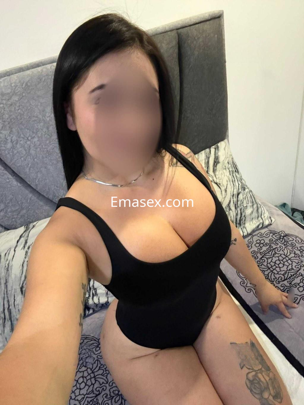 Escort en Barcelona posando en una cama con un body negro, cabello largo y lacio, y tatuajes visibles en el brazo.
