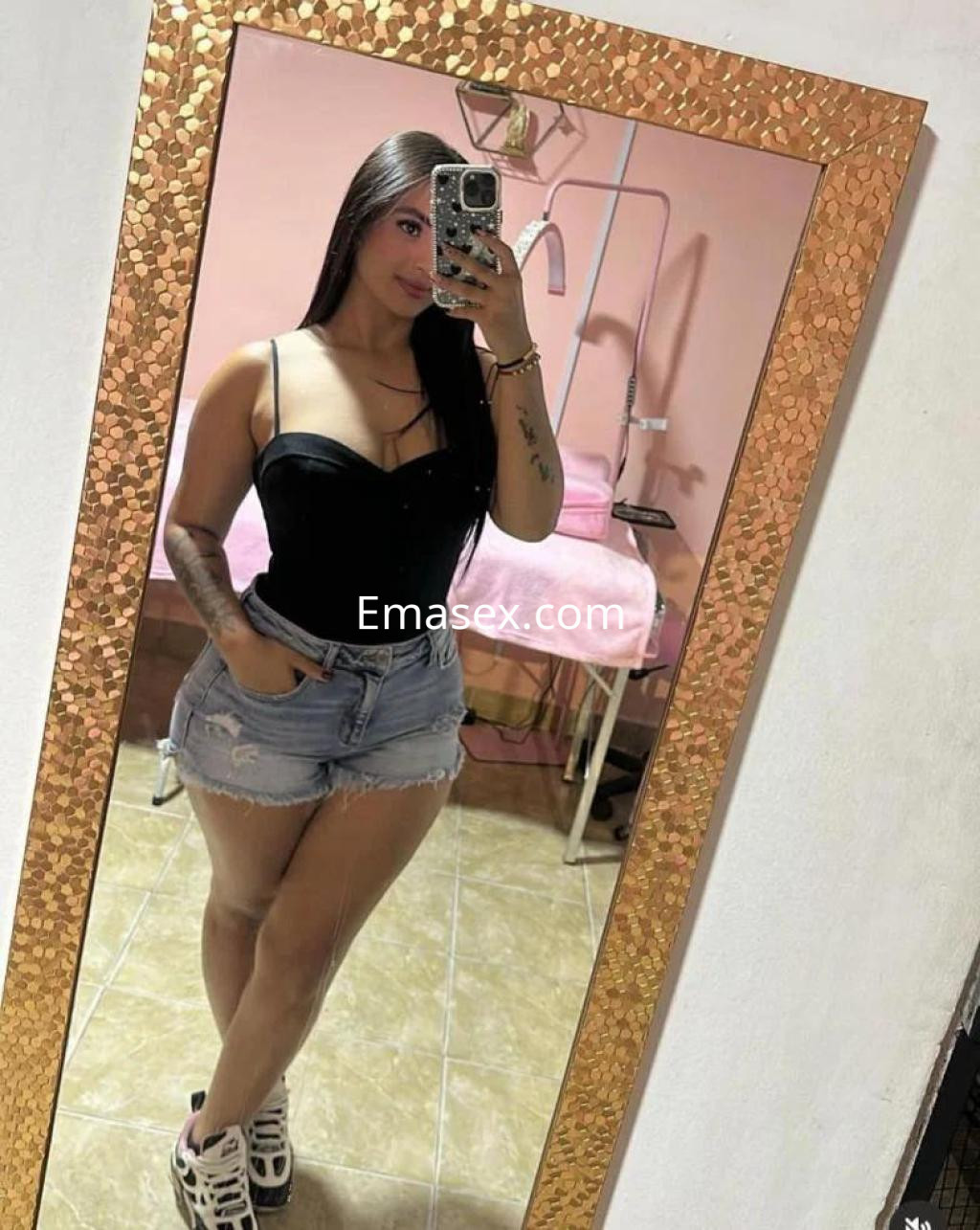 La puta en Cáceres se muestra en un espejo, luciendo un top negro y shorts de mezclilla, con zapatillas deportivas.