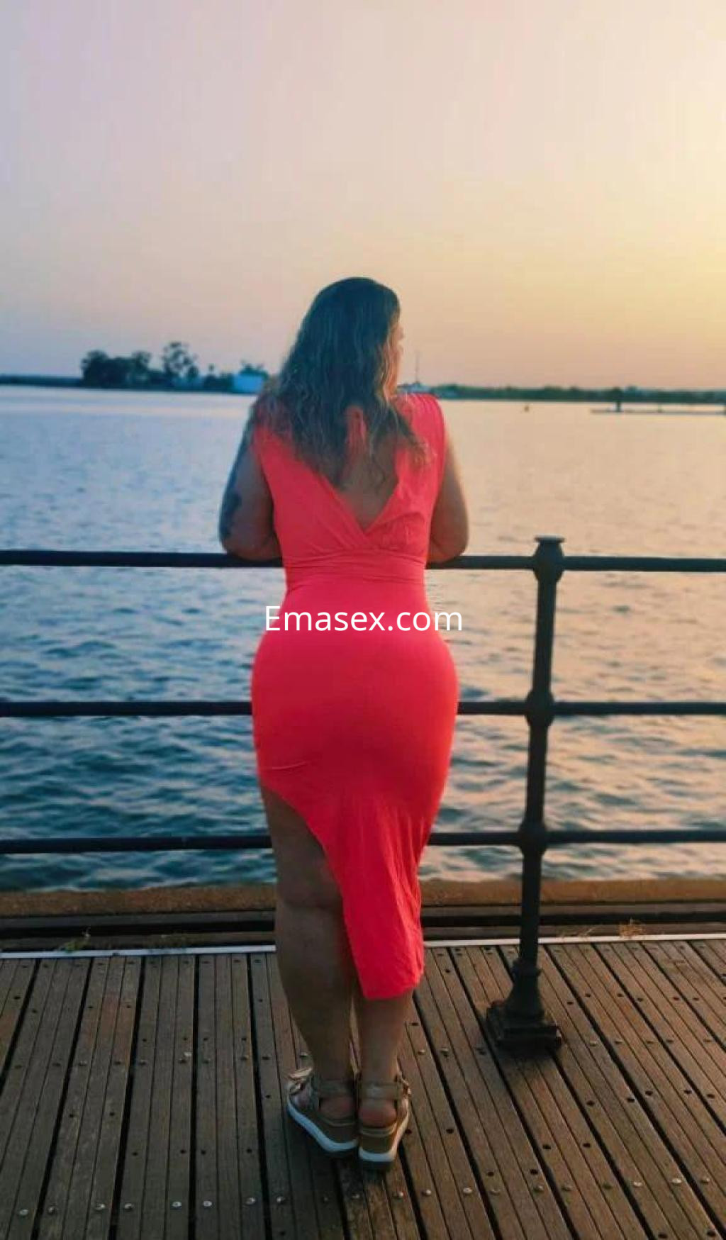 Puta de pie junto al agua en Huelva, luciendo un vestido rosa con una abertura y cabello ondulado.