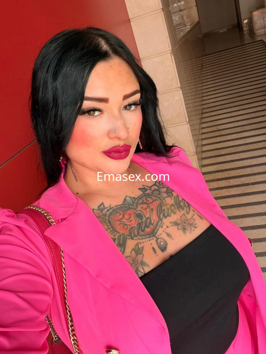 Puta en Madrid con cabello largo y oscuro, vistiendo un conjunto rosa y blusa negra, mostrando tatuajes en el pecho.