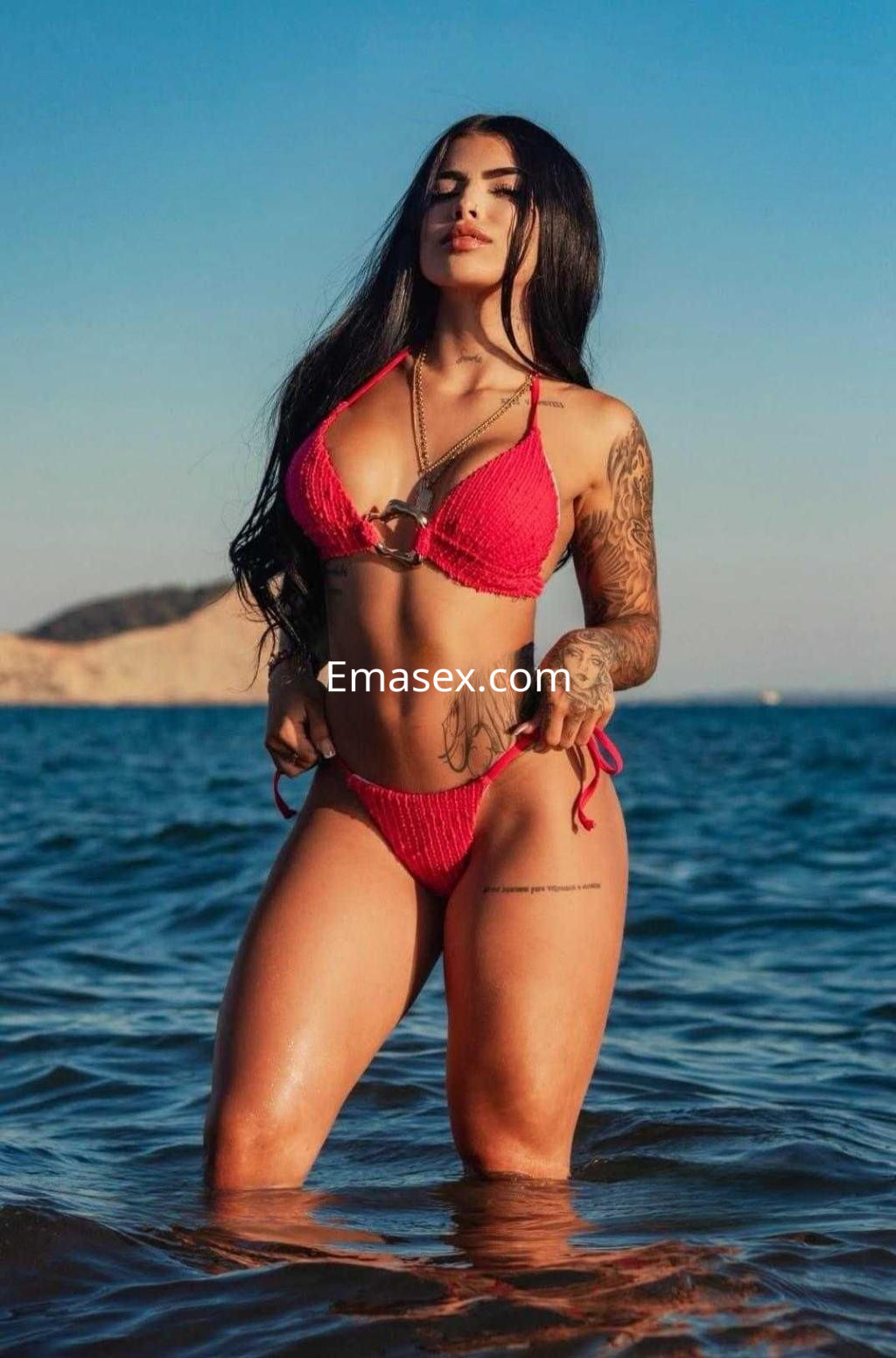 Puta en Ibiza luciendo un bikini rojo mientras posa en el agua con el sol de fondo y cabello largo.