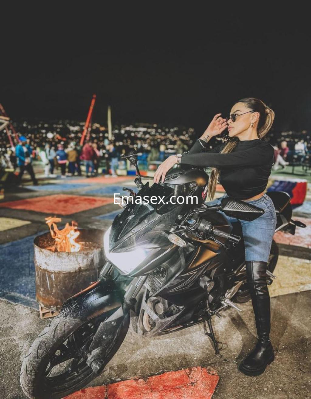Escort posando sobre una moto negra, vestida con un suéter negro y leggings de cuero, en un entorno nocturno en Girona.