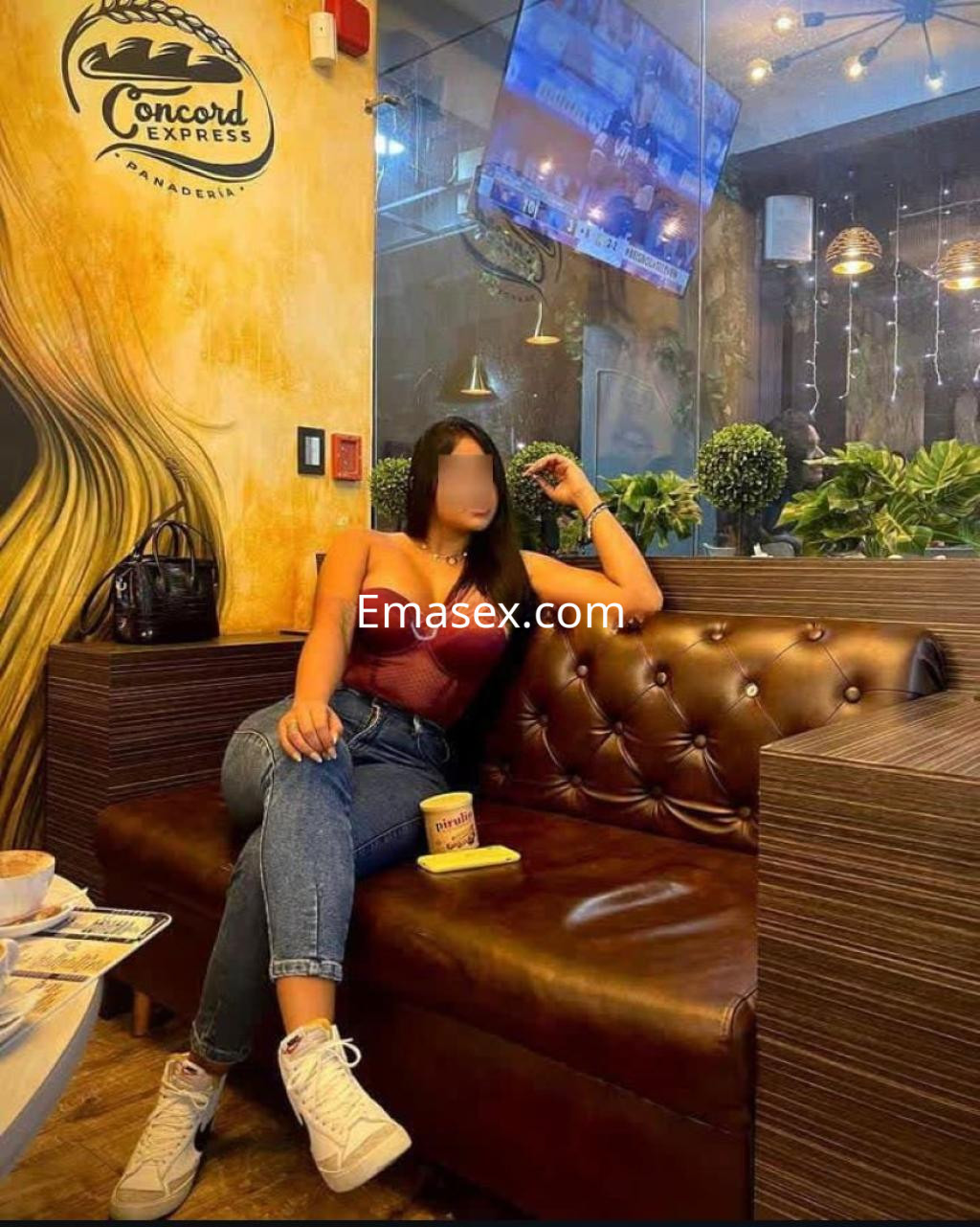 Escort sentada en un sofá marrón, vistiendo un top rojo y jeans, en un ambiente moderno de Zaragoza.