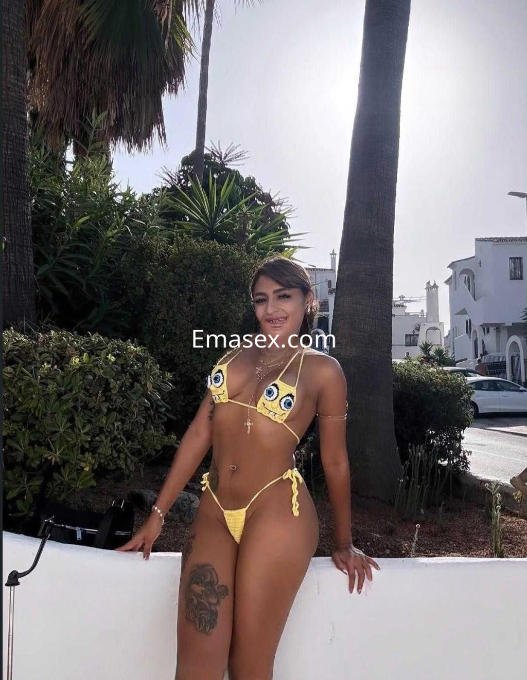 Escort en Granada, con bikini amarillo y detalles coloridos, posando en un entorno al aire libre rodeada de palmeras.