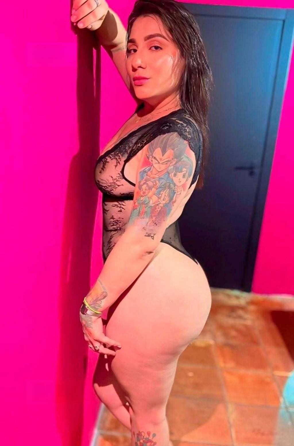 Puta con cabello oscuro y largo, usando un body negro, posa de perfil contra una pared rosa en Zaragoza.