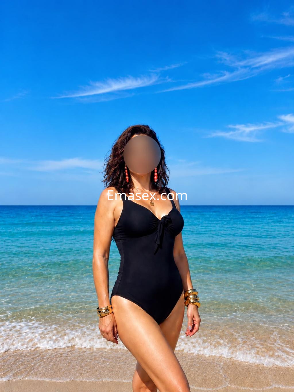 Escort en Vitoria vistiendo un bañador negro, posando en la playa con el mar y el cielo azul de fondo.