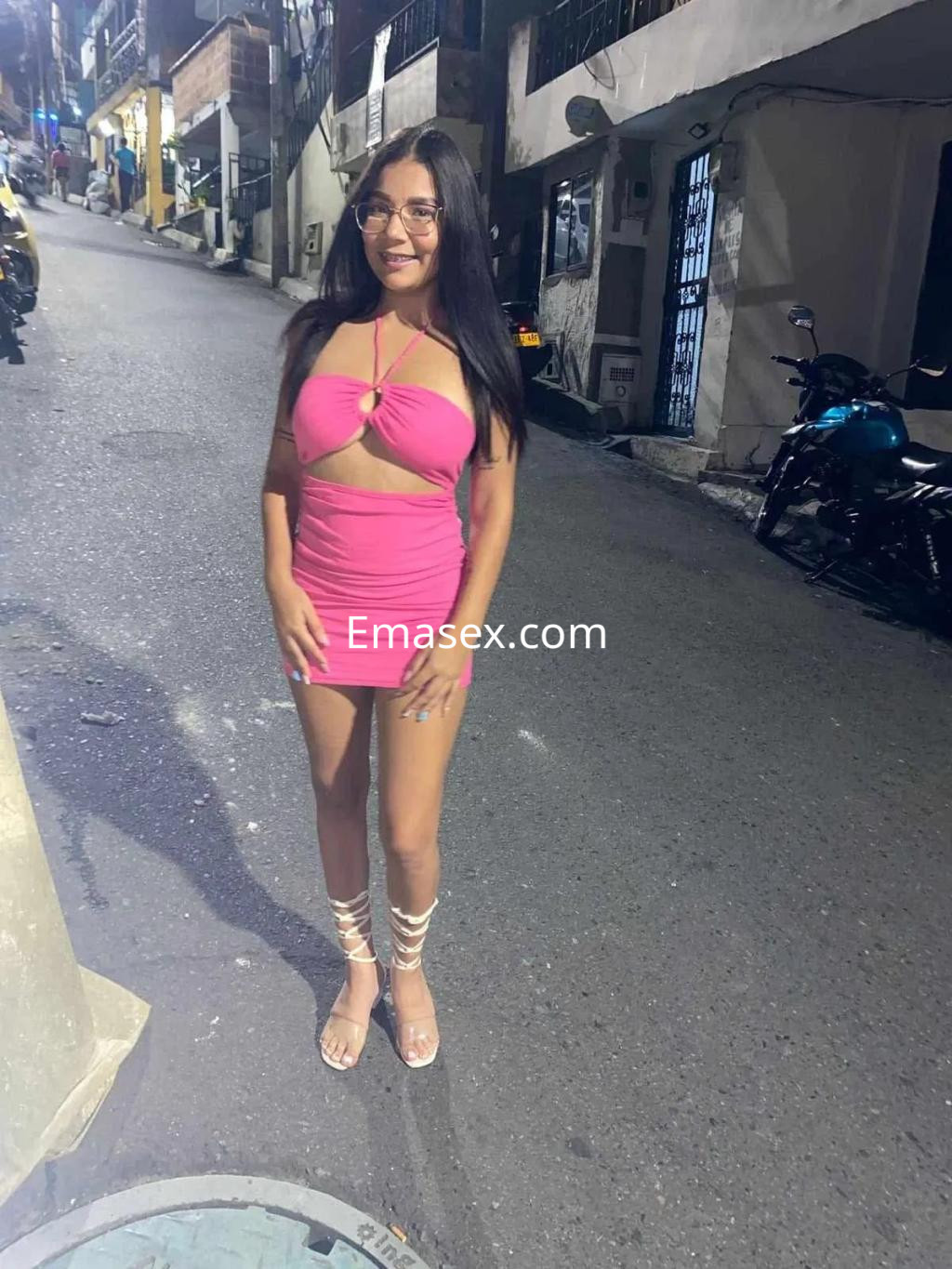 Puta de cabello largo y lacio, viste un vestido rosa ajustado y sandalias, posando en una calle de Oviedo.