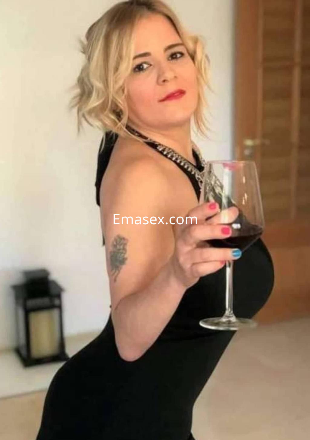 Puta en Zaragoza luciendo un vestido negro ajustado, sosteniendo una copa de vino y con cabello rubio ondulado.