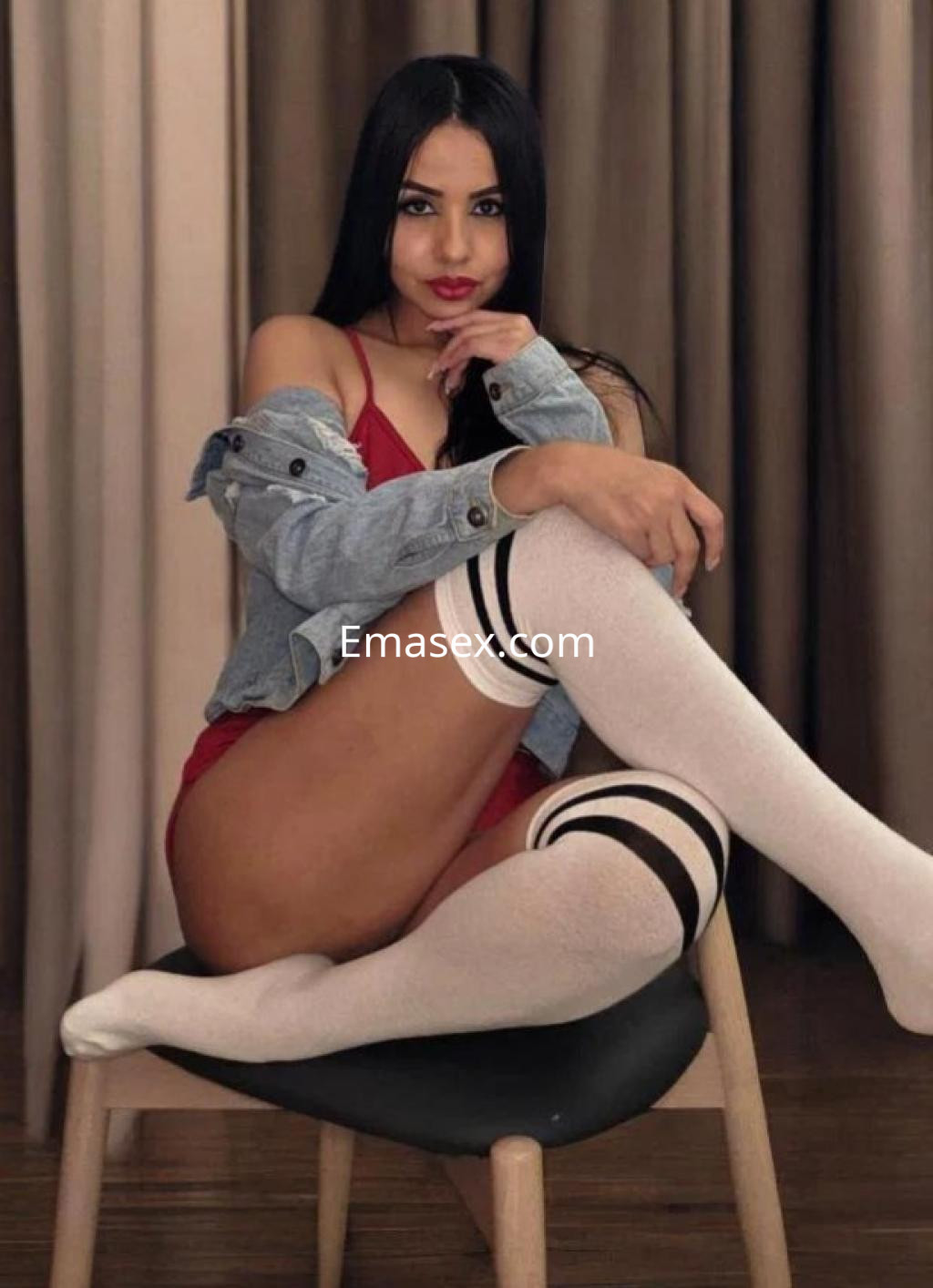 Escort en Las Palmas, sentada en una silla con un body rojo, chaqueta de mezclilla y medias largas blancas, luciendo coqueta.