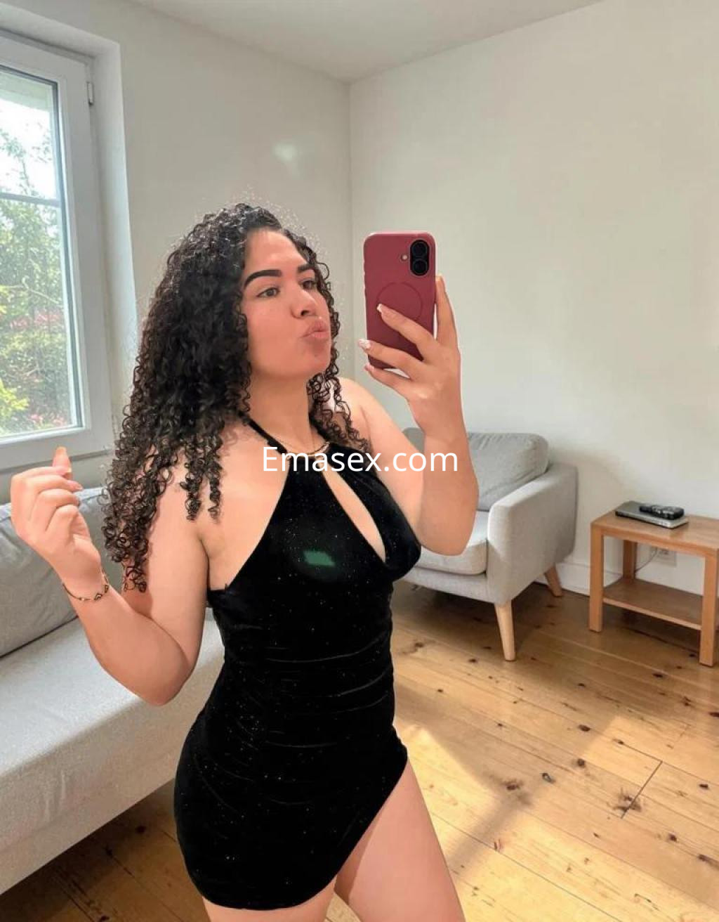 Escort en Barcelona posando con un vestido negro ajustado y cabello rizado, mientras se toma una selfie en un ambiente luminoso.