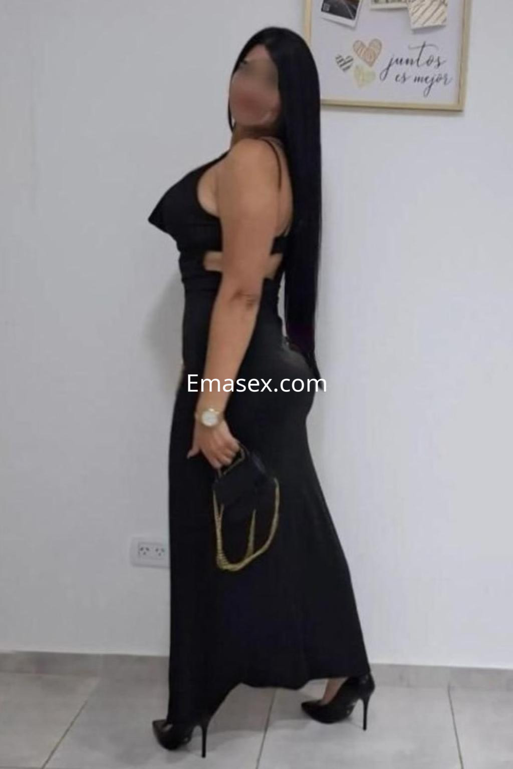 La puta viste un elegante vestido negro ajustado y tacones altos, posando de lado en un ambiente luminoso en Madrid.