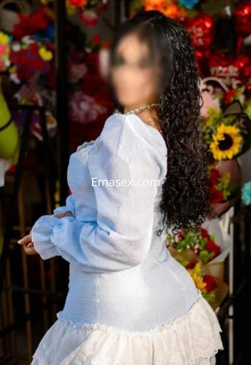 Puta en Santander lleva una blusa de manga larga azul claro y una falda blanca, con un fondo colorido de flores.