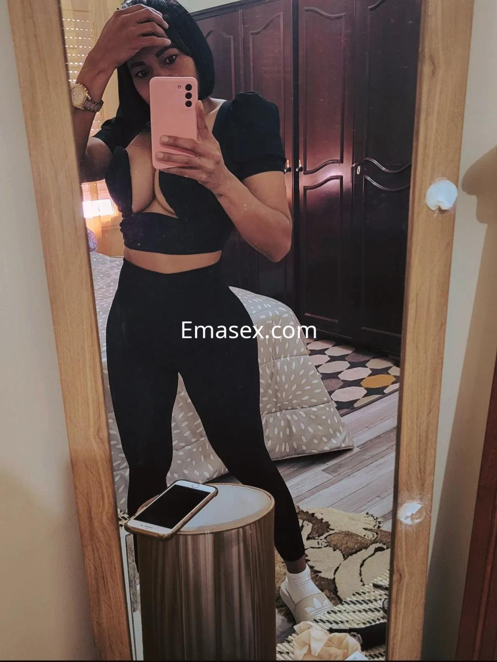 Escort en Valladolid con blusa oscura de escote y pantalones ajustados, posando frente a un espejo en un dormitorio.
