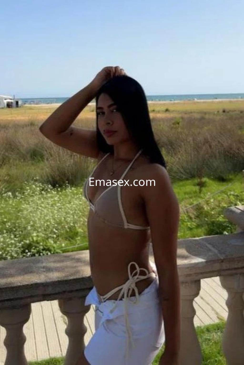 Puta en Barcelona, usando un bikini beige y una falda blanca, posando con el mar y la naturaleza de fondo.