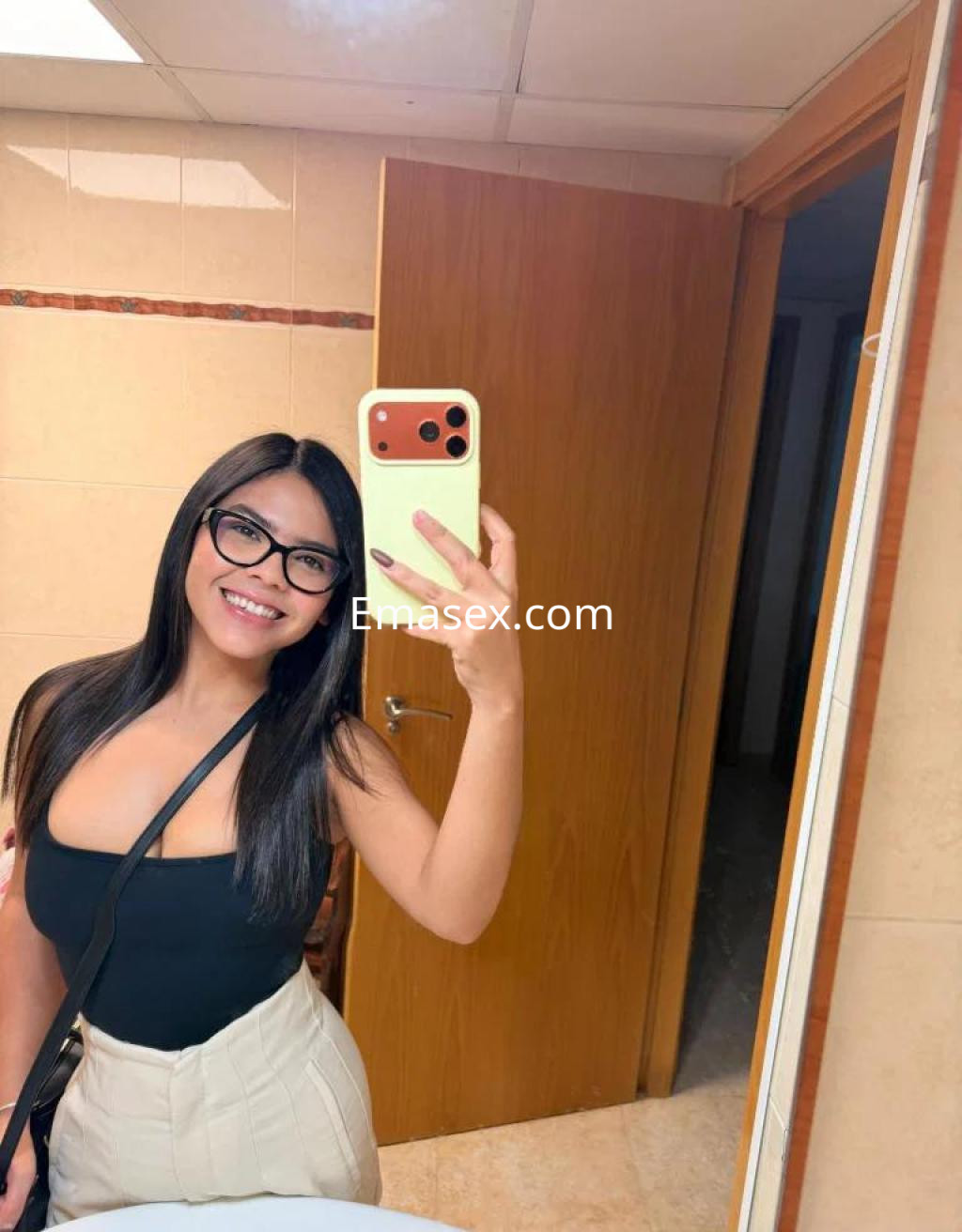 La puta en Tarragona sonríe mientras se toma una selfie en un baño, vistiendo un top negro y pantalones claros.