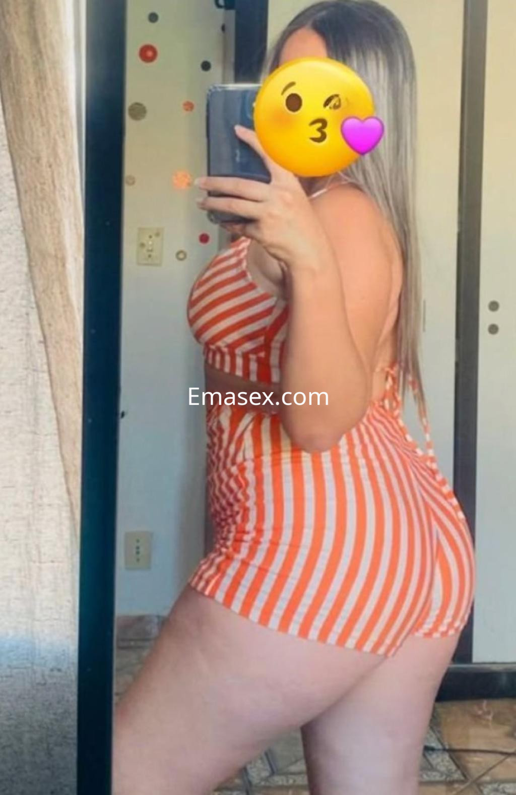 Puta en Cádiz posando de perfil frente a un espejo, con un conjunto de baño a rayas naranjas y blancas.
