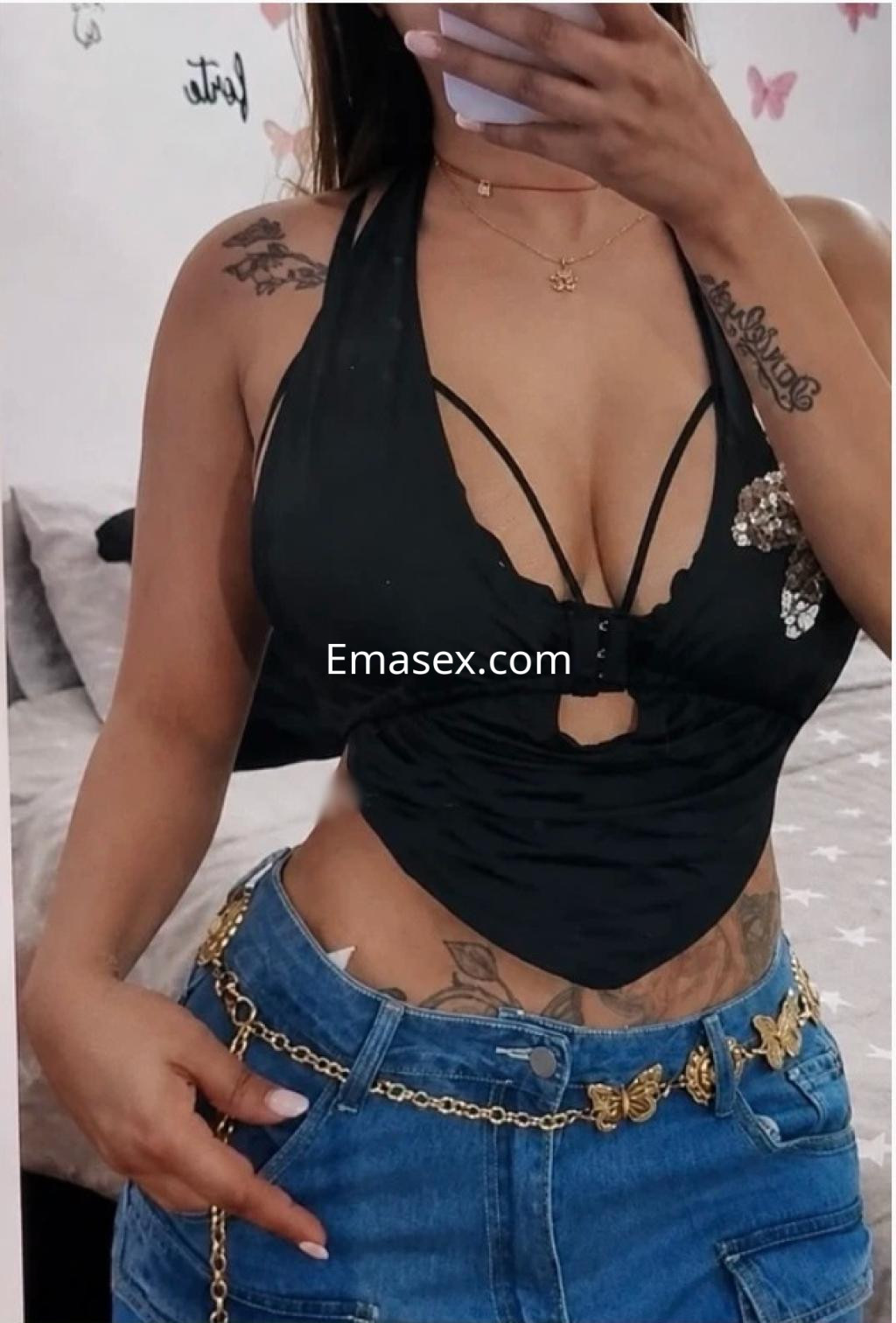 Puta en Gijón viste una blusa negra con detalles en el escote y jeans ajustados, mostrando sus tatuajes.
