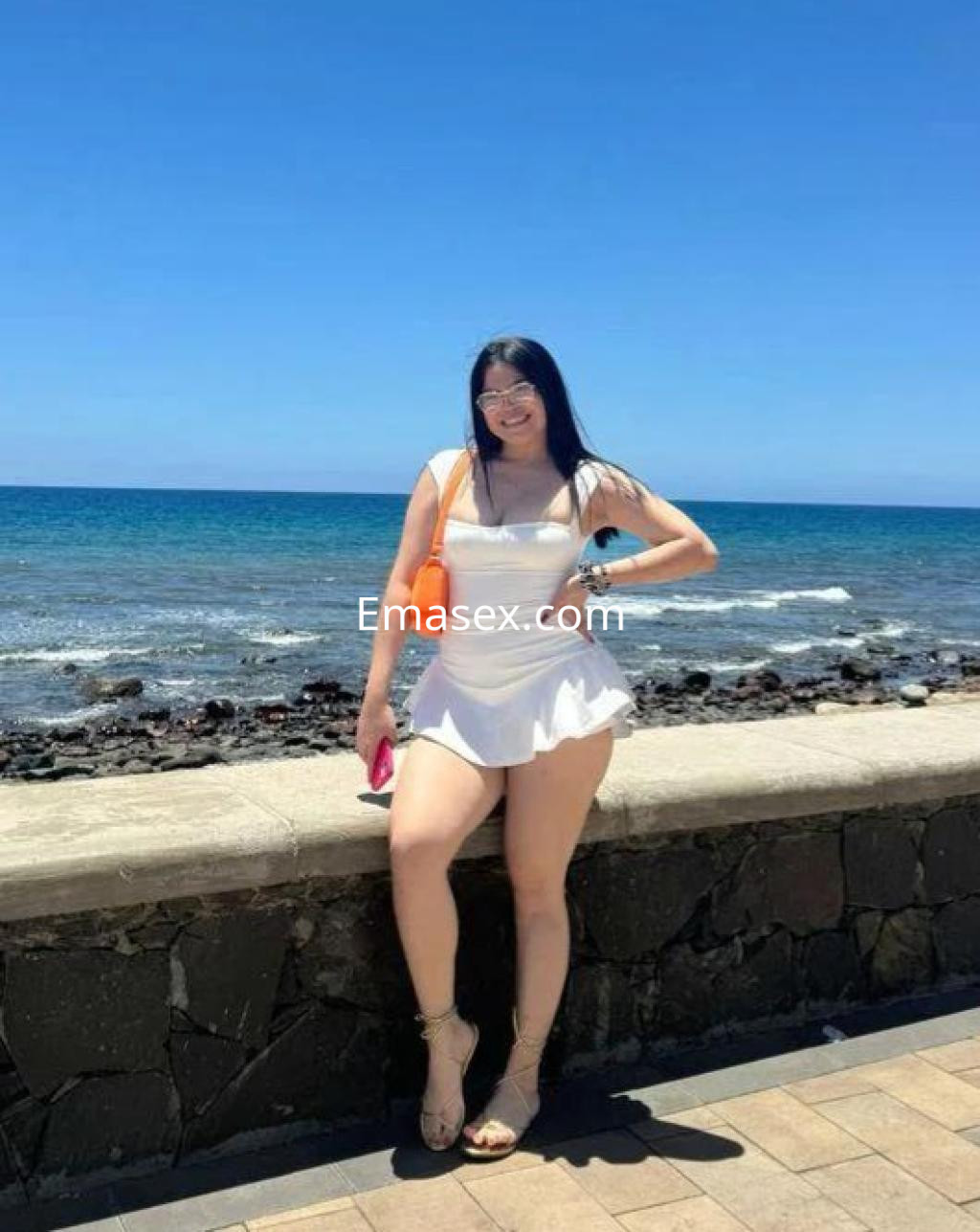 Puta vestida con un vestido blanco corto y sandalias, posando junto al mar en Tenerife bajo un cielo despejado.