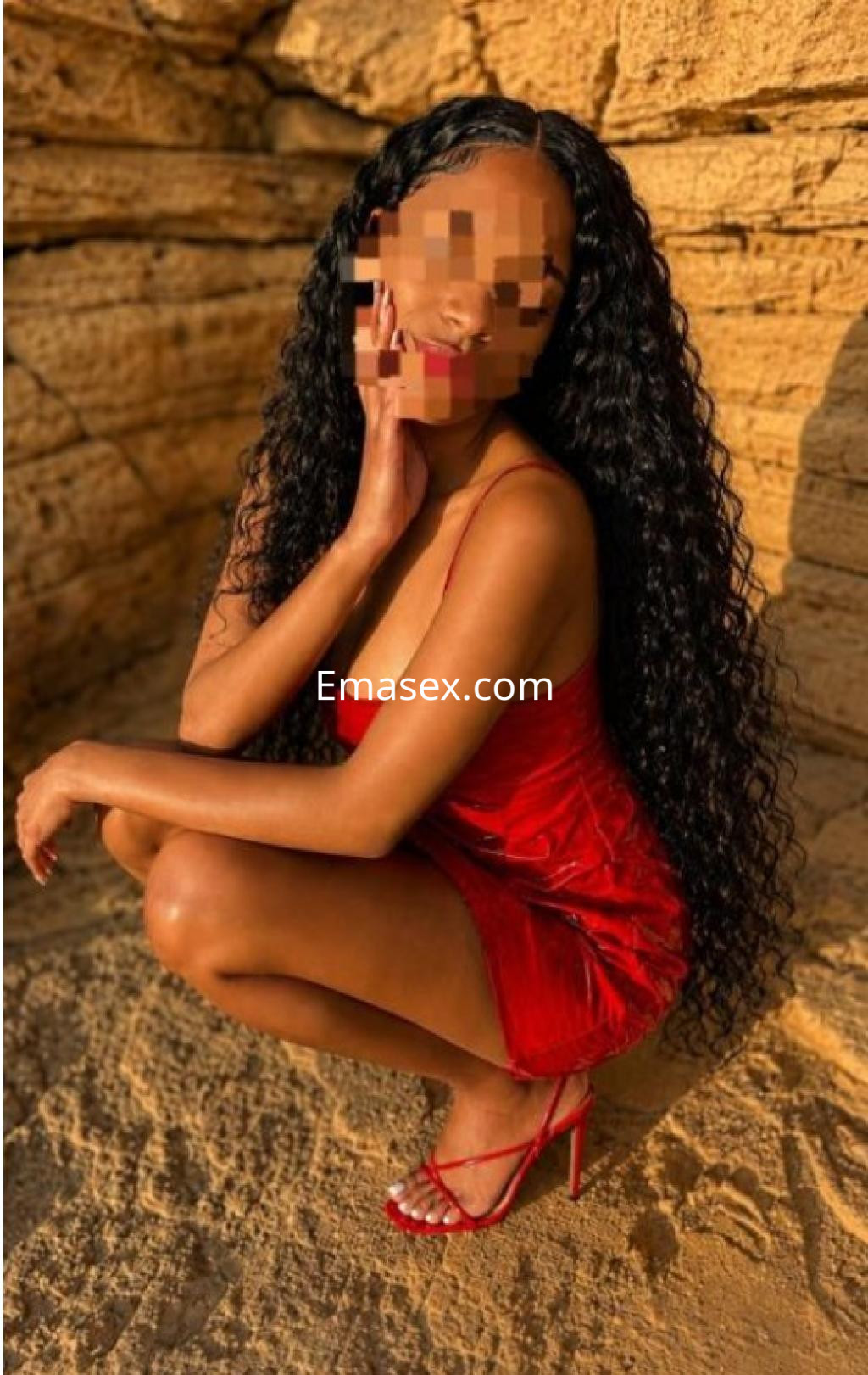 Escort en Gijón, con vestido rojo y sandalias, posando de cuclillas sobre una superficie arenosa.