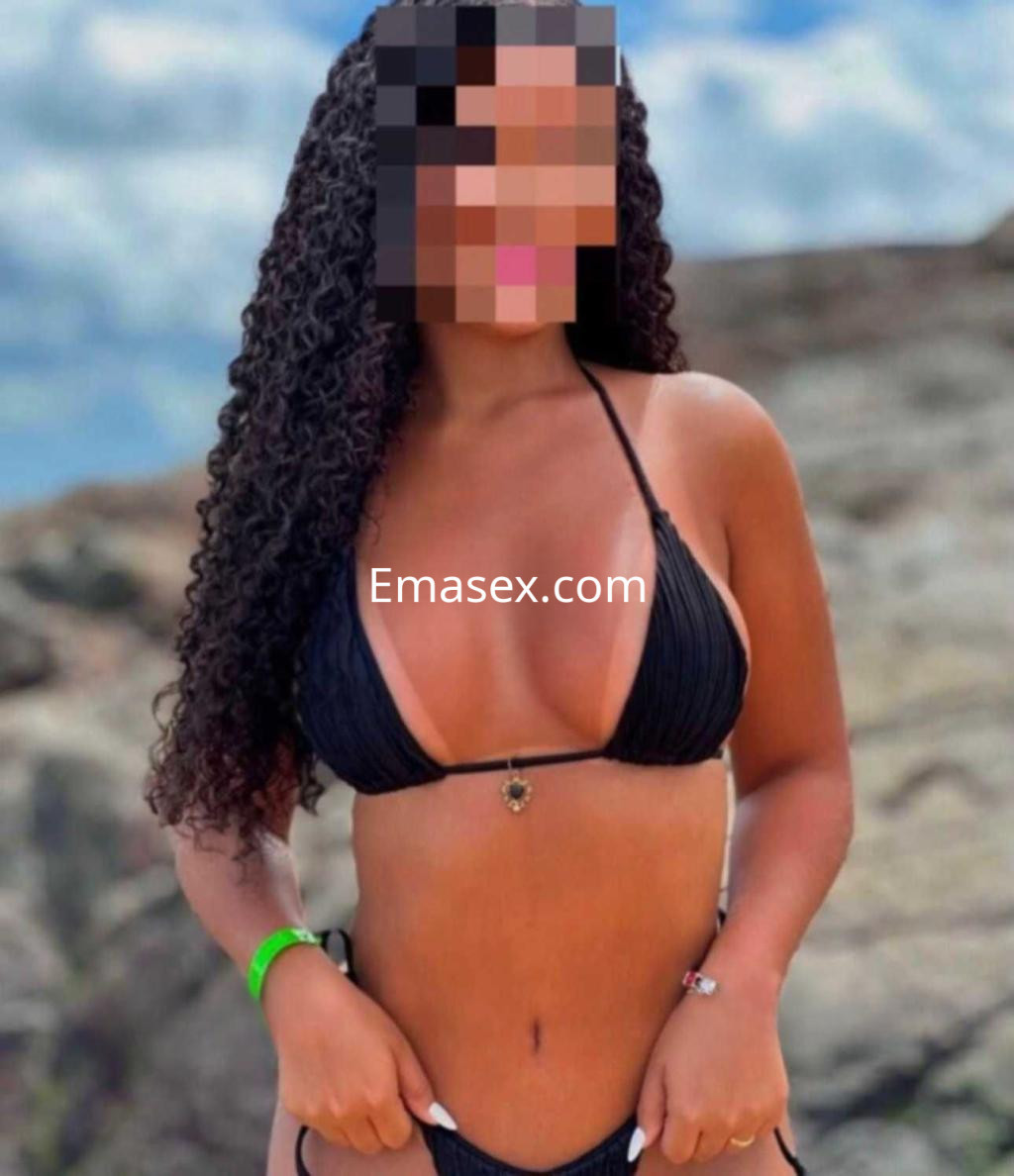 Puta de cabello rizado y largo, luciendo un bikini negro en una playa rocosa bajo un cielo despejado en Zaragoza.