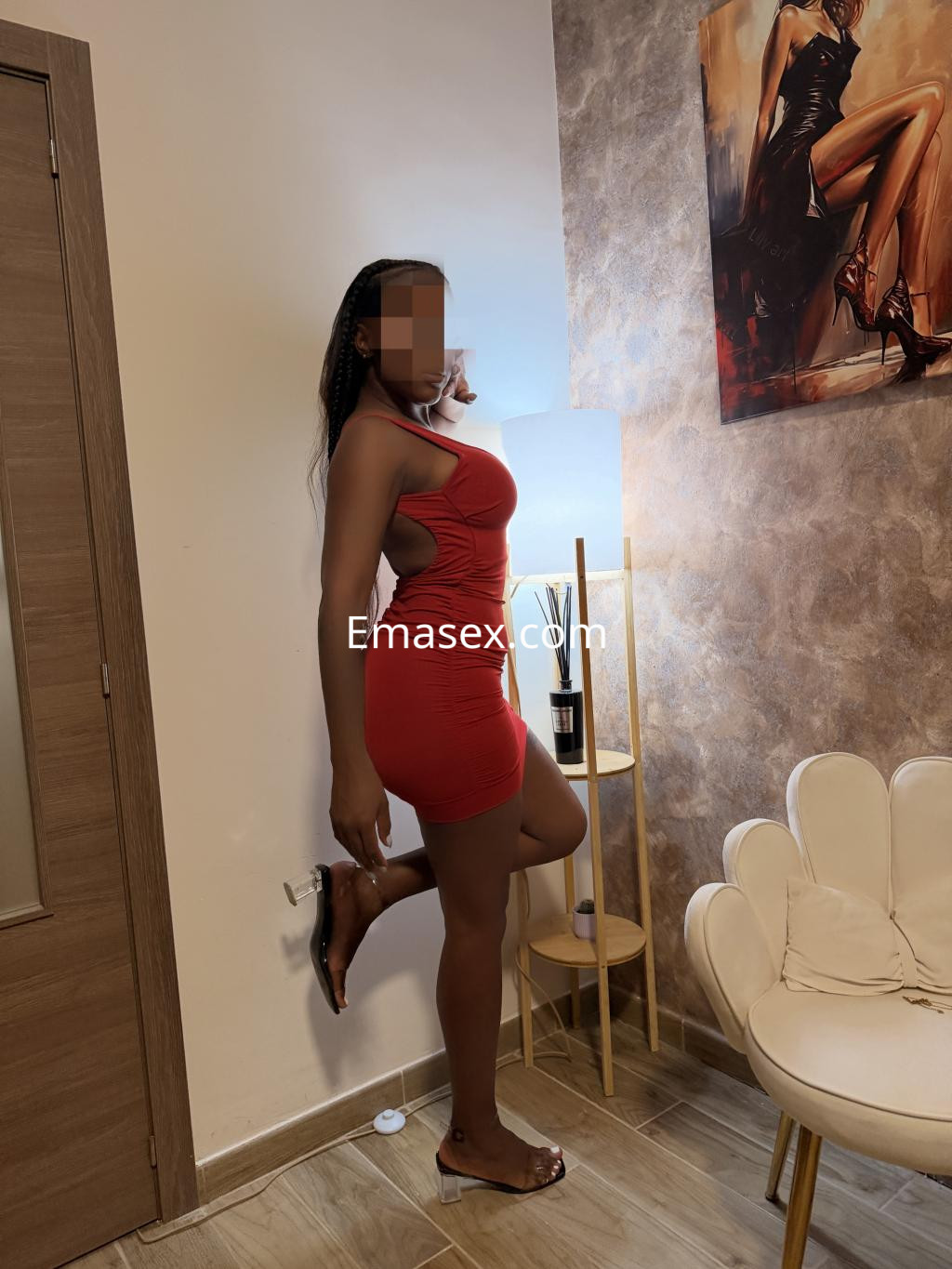 Escort en Madrid posando junto a una lámpara, luciendo un vestido rojo ajustado y sandalias transparentes.