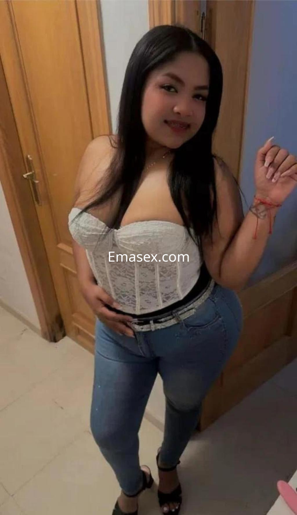 Escort en Cádiz viste un corsé blanco y jeans ajustados, posando con una mano en la cadera y una sonrisa.