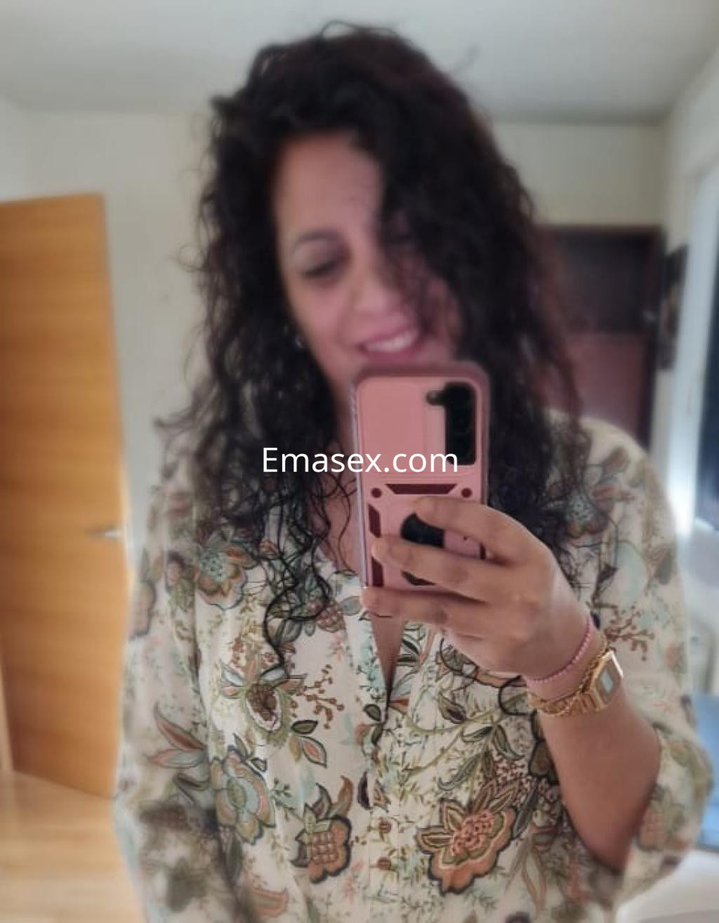 Escort en Pamplona, con cabello rizado y blusa estampada, sonriendo mientras se toma una selfie en un entorno interior.