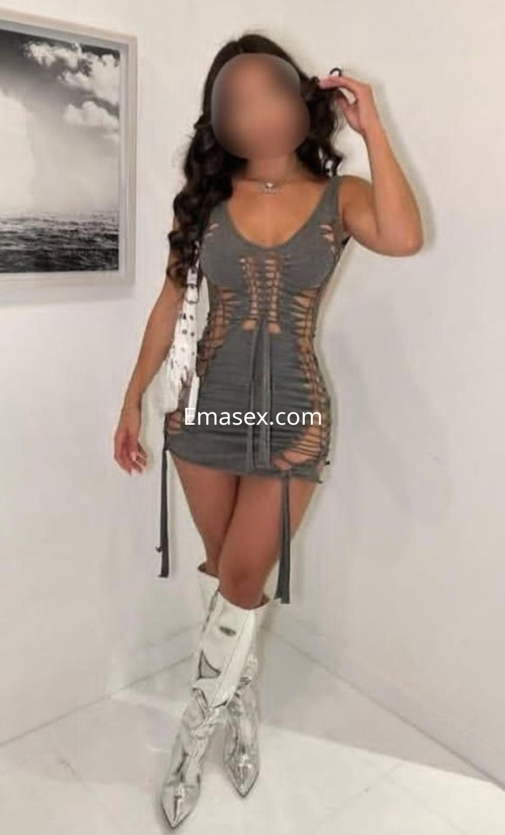 Escort en Barcelona, lleva un vestido gris con cortes laterales y botas plateadas, posando en un fondo claro.