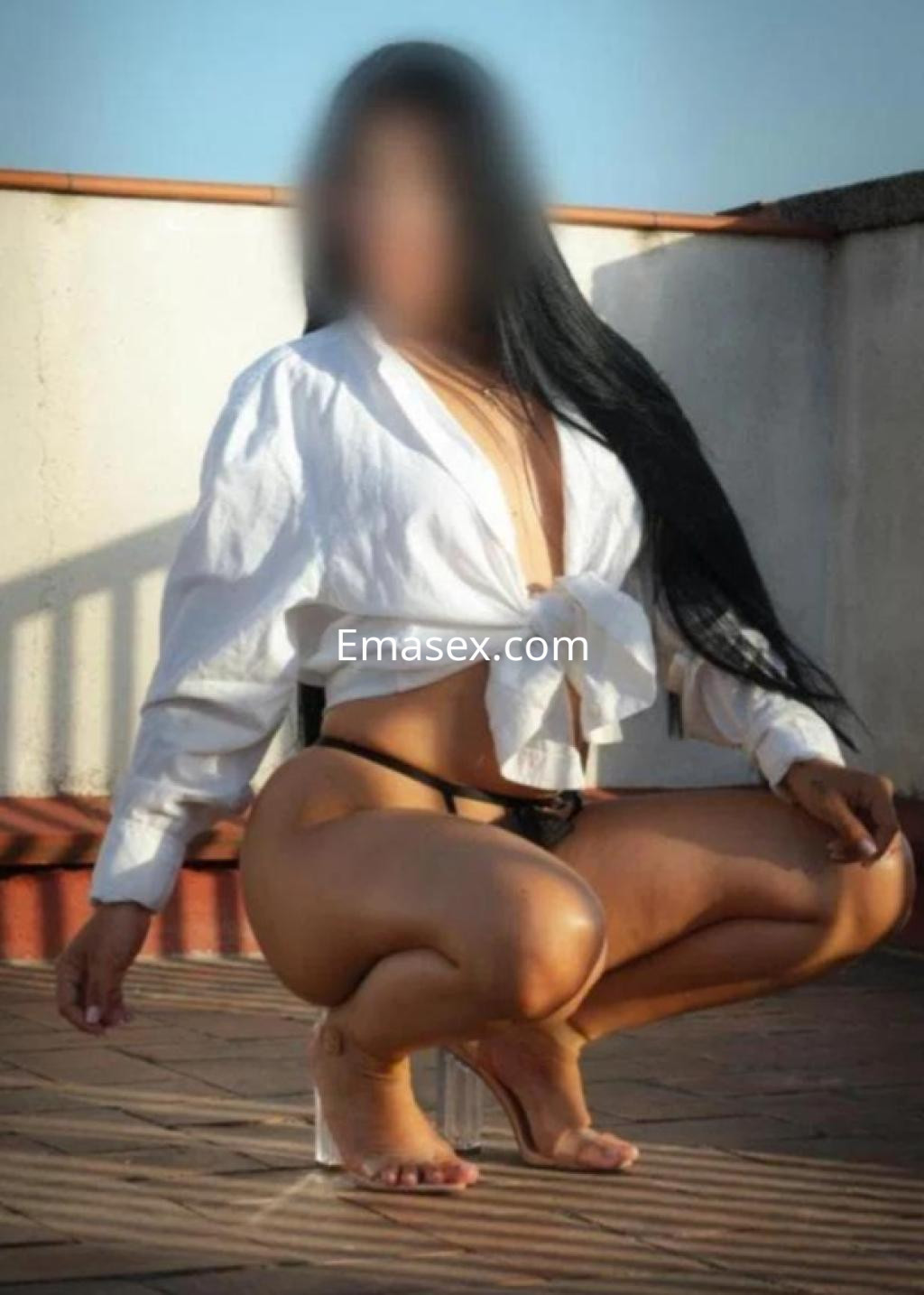Escort en Barcelona con cabello largo y lacio, viste una blusa blanca y una tanga negra, posando en una terraza.