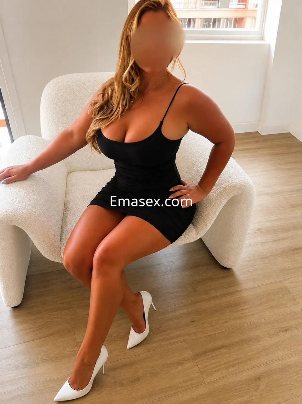 Puta en Palma de Mallorca, sentada sobre un sillón blanco, vistiendo un vestido negro ajustado y tacones blancos.
