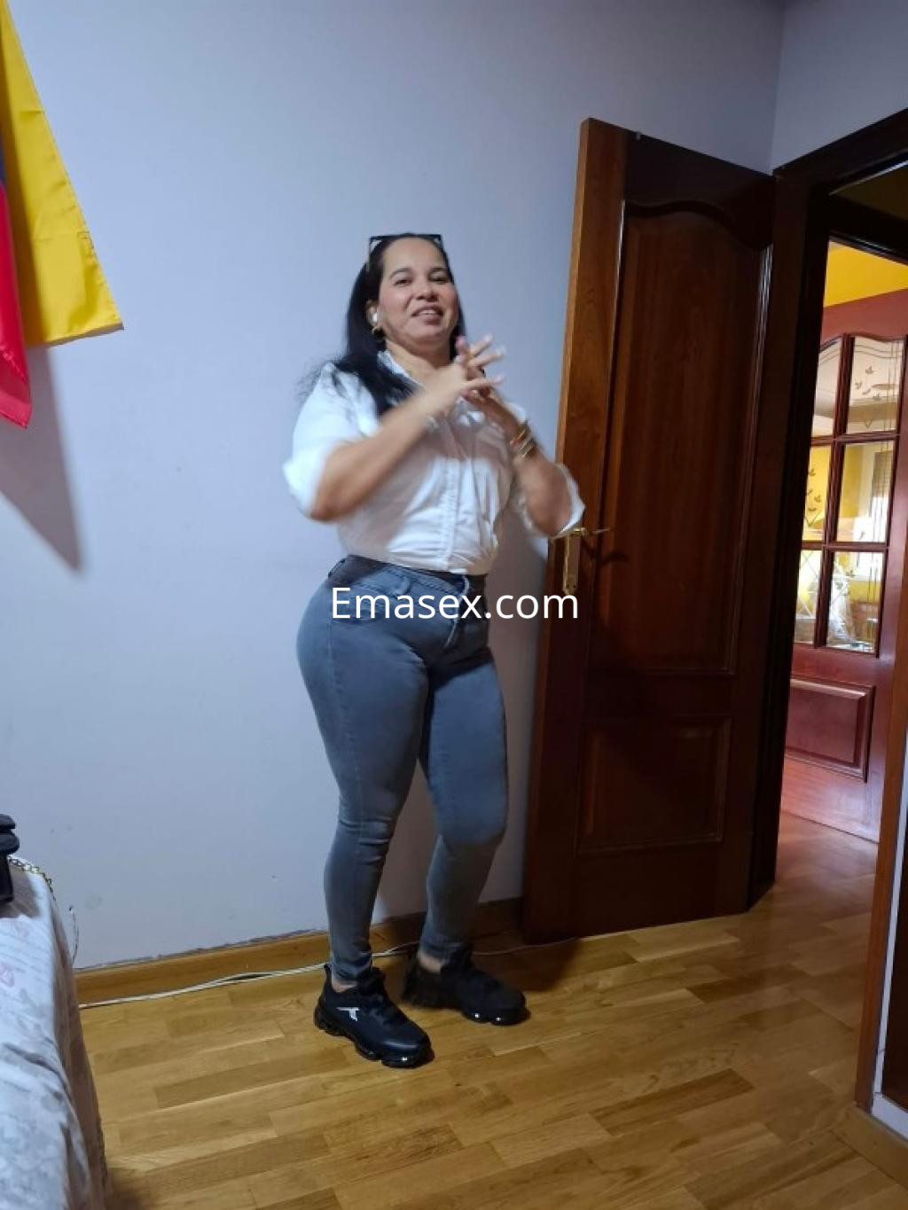 Puta en Oviedo luce una blusa blanca y jeans ajustados, posando con una sonrisa en un interior acogedor.