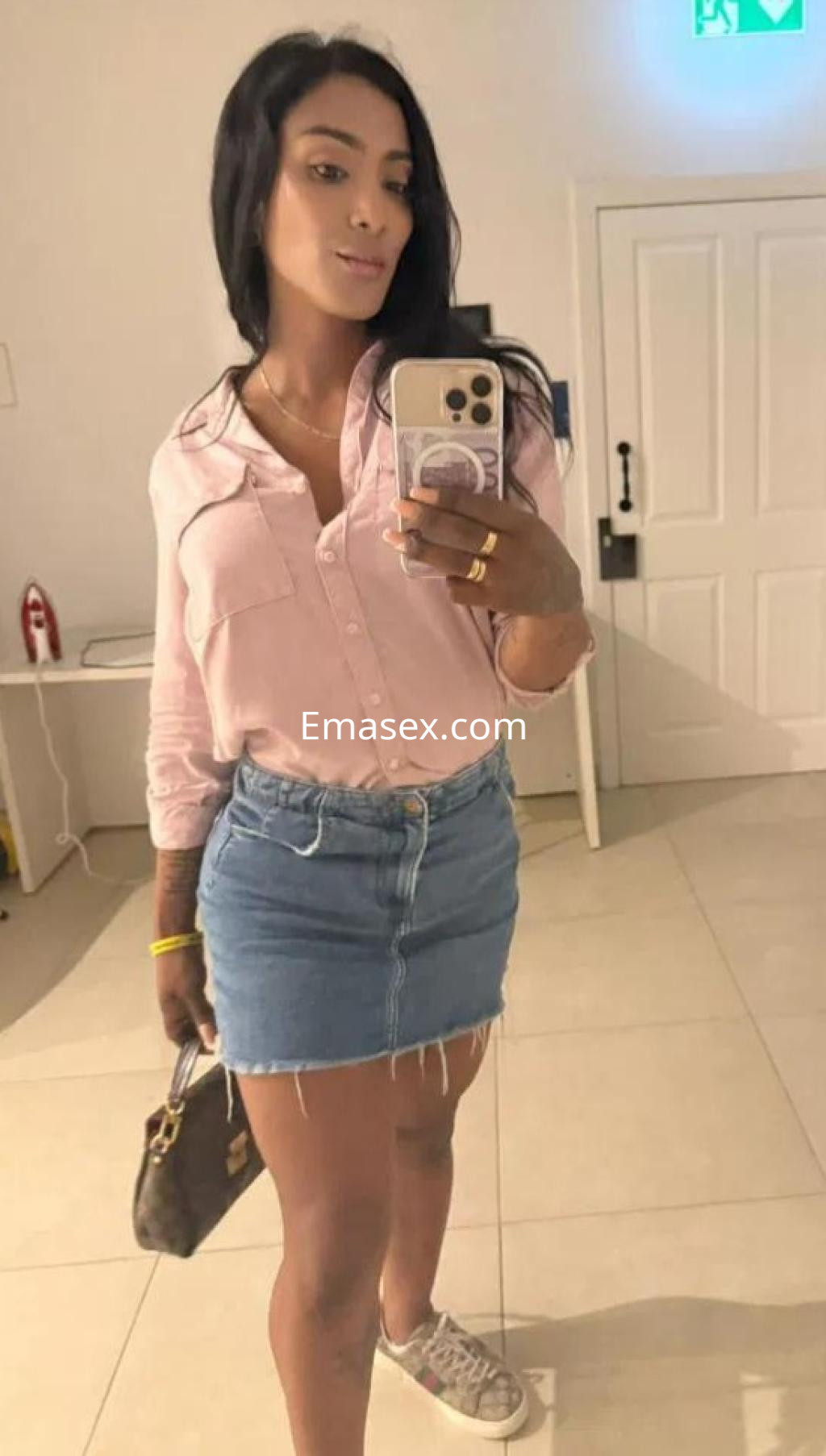 Puta en Barcelona posando en un espejo, con camisa rosa y falda de mezclilla, luciendo un estilo casual.