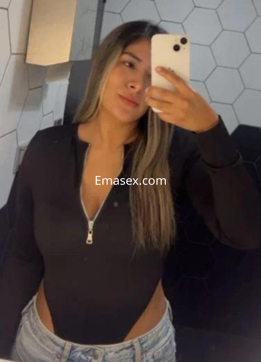 La escort en Barcelona se toma una selfie en el baño, luciendo un body negro y con el cabello largo y lacio.