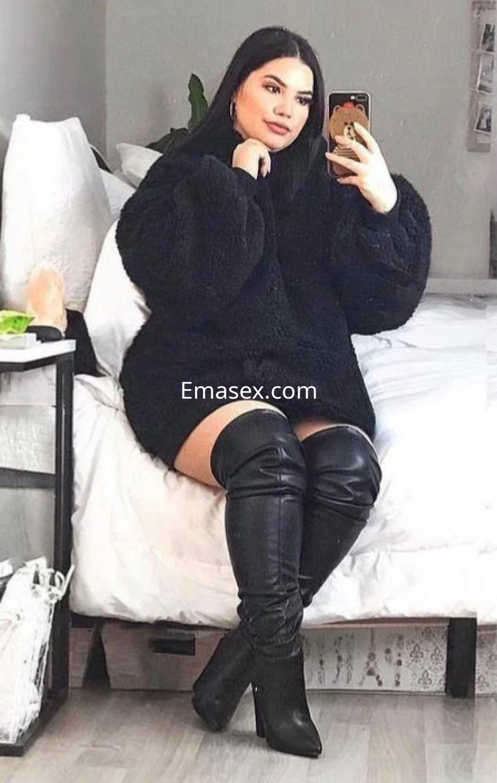 Escort en Barcelona sentada en una cama, vistiendo un abrigo negro y botas altas, mientras se toma una selfie.